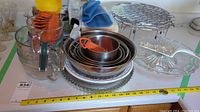 Overview of various cooking bowls, measuring jugs, plastic utensils, metal baking pans, glass serving dish, and cooling rack arranged on a countertop with a measuring tape for scale.