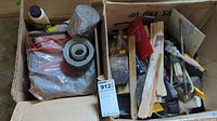 Two cardboard boxes filled with assorted hardware items such as lubricants, sanding blocks, and paint brushes.