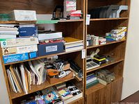 Wooden shelving filled with assorted office supplies including folders, boxes of staples, files, desktop accessories, calculators, and sorters.