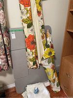 Folding sewing table leaning against the wall with floral fabric rolls and some white fabric pieces piled at the base. Hooks and blue storage bag visible.