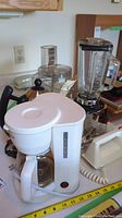 Photo showing several small kitchen appliances including a white drip coffee maker with glass carafe, a stainless steel electric kettle, a blender base with clear pitcher, and a food processor bowl.