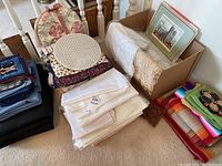 Photo shows stacked piles of tablecloths, folded placemats, a cardboard box with additional linens, and framed picture on top