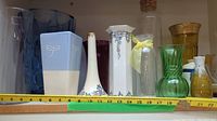 Assorted glass and ceramic vases and glasses arranged side by side with measuring tape showing sizes ranging from about 2 inches to 15 inches.