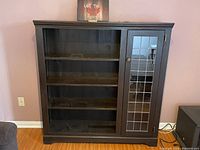 Front view of the wooden cabinet showing three open shelves on left and the leaded glass door on the right side with knob and metal hinges. Black finish with crown molding top.