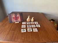 Photo of 10 hand-painted ceramic herb tiles with botanical names in cursive and two small beige ceramic vases on wood table.