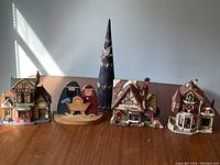 Overview of the set showing four ceramic Christmas village houses, blue ceramic Christmas tree, and wooden nativity scene on a wooden surface near a wall with lighting from a window.