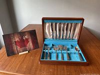 Stainless steel cutlery set with knives arranged in upper compartment and other utensils in lower compartment of wooden box with blue and white fabric lining.