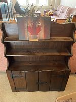 Frontal view of dark stained folk art wooden shelf showing three open shelves and lower cabinet with three doors secured with latch and iron hinges.