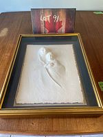 Framed embossed print visible within a shadow box frame with gold outer trim and black matte interior, resting on a table with a wooden texture and an auction lot card in the background.