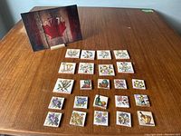 Overview of 25 square decorative ceramic tiles with botanical, fruit, and kitchen themes arranged on a wooden table.