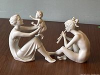 Side view of both white matte porcelain figurines on wooden surface, showing woman with child and nude girl playing flute