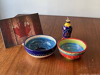 Three items on a wooden table: two ceramic bowls, one blue with black and white designs, one multicolored with blue, green, red; accompanied by a blue and yellow ceramic oil jar with cork stopper.