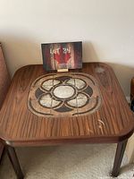 Front view of the side table showing square wooden top with tile inlay in concentric floral pattern, some surface scratches visible.