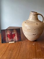 Photo of jug-shaped floor vase with handle, showing full profile, placed on wood surface against plain wall.