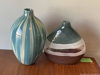 Pair of mid-century modern glass vases: taller teal and white striped alongside shorter rounded multi-colored vase on wooden surface.