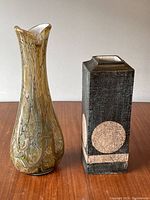 Front view of two vases on a wooden surface showing a tall swirl-pattern glass vase and a rectangular pottery vase with geometric designs.