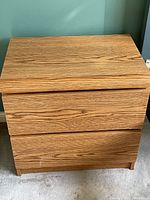 Front view of rectangular night table with two drawers, oak wood grain veneer, placed on carpeted floor against green wall.