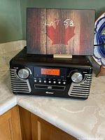 Front view of the black Memorex retro portable turntable audio system displaying AM/FM frequency 107.9 on the digital screen with volume and tuning knobs and speaker grilles on either side.