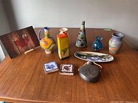 A set of nine pottery and ceramic decorative pieces displayed on a wooden table with varying shapes and colorful finishes including a conical vase and a flask with a Mayan motif.