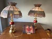 Two vintage stained glass table lamps displayed side by side on wooden table. Both lamps feature scalloped stained glass shades with pastel colors. Bases are brass with a polished circular design and a red spherical accent. One shade missing one piece and has a crack on top.