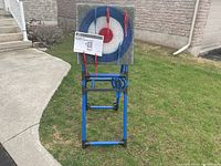 Photo shows the assembled blue metal frame stand with the target and axes held on it, placed outdoors on grass near sidewalk and house wall.