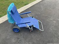 Blue fabric beach chair with metal frame on outdoor pavement, two large rear wheels visible, fabric sun shade attached.