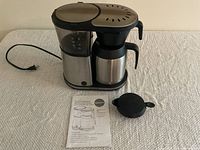 Bonavita 8 cup coffee brewer with black and stainless steel finish, lid, and owner's manual on table.