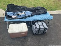 Single portable bed frame with blue air mattress and black cover, Coleman Duramate cooler and Hike Crew portable propane water heater in gray soft case, shown on pavement with grass background