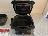 Close-up view of open Ninja Foodi Grill with square cooking basket positioned inside and control panel visible on front face of unit.