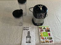 Full overview of blender base, two blending containers, user manual, and recipe brochure on a carpeted surface.