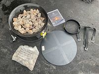 Outland propane firebowl bowl filled with lava rocks, including extra lava rocks in a bag, metal cover, and propane tank hold-down straps on black asphalt surface.