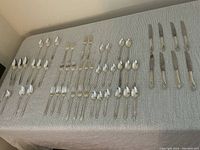 Full spread of all flatware items laid out on a table cloth showing the number and variety of pieces including forks, knives, various spoons, and salad forks.