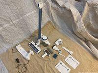 Full layout photo of the vacuum, multiple attachments, dock station, power adapter, manual booklets on beige surface