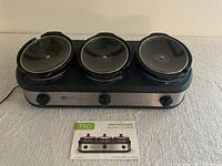 Full front view of TRU Three Crock Buffet Slow Cooker with three black crock pots and glass lids, showing the front control panel with knobs and TRU branding. The owner's manual is placed in front of the cooker.