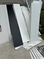 Photo showing several white and one black long shelves lying against railing outdoors, part of shelving lot.