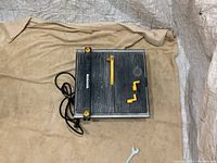 Top down view of the Mastercraft wet tile saw, showing the rectangular cutting surface with grooved black platform, yellow adjustable guide, yellow clamp accessory attached, and the power cord wrapped to one side, plus a wrench placed on the cloth.