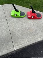 Two Zipfy luge seats, green and red, shown from a three-quarter side view on concrete sidewalk outside, positioned next to grass and driveway edge.