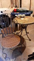 Photo showing round mosaic stone top table and one wicker seat cast iron chair from angle revealing table leg design and chair seat.