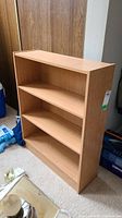 Full view of the laminate bookcase shelf showing front and side, placed on carpet with visible wear on surfaces.