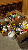 Wide view showing multiple cardboard boxes filled with assorted cleaning bottles including spray nozzles and caps.