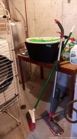 Photo showing black and white storage bins, broom and spray mop, drying rack, and cleaning products on a wooden table and floor in basement.
