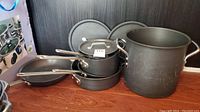Photo showing large stockpot, saucepans with lids, and fry pans grouped together on floor against black wall.