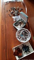 Photo of assorted costume jewelry including rings on a glass ring dish, brooches in box, pierced earrings scattered on wood surface.