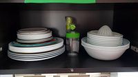 Shelf showing stacked ceramic plates with different colored rims, white bowls, a white plastic citrus juicer on top of bowls, and two glass dispensers with green parts.