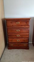 Front view of wooden dresser with five drawers and brass handles on a beige carpeted floor against a white wall.