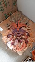 Large handcrafted feather face mask displayed on a cardboard surface showing the full length of feathers arranged in layers.