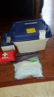 Photo of a plastic storage bin with blue lid, a small red first aid kit pouch, and a clear bag of surgical masks on tiled floor