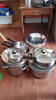 Photo showing stacked Verona stainless steel pots and pans with glass lids and black handles on a wooden floor.