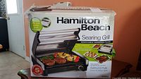 Front of box showing Hamilton Beach searing grill with food inside grill and key features such as removable nonstick plate.