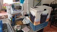 Mainstay paper shredder in box, assorted office and cleaning supplies on table, including storage bins, pens, hand soap, coaster, and index cards.